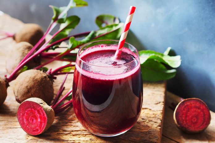 Copo de vidro com suco de beterraba e um canudo. Ao fundo, várias beterrabas cortadas. Metrópoles