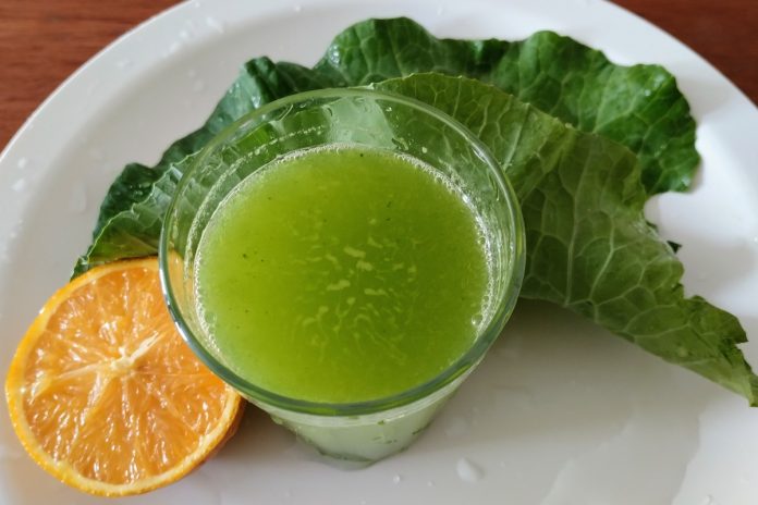 Foto mostra suco verde, suco de laranja com couve. Em um prato branco estão dispostos ambos ingredientes ao lado de um copo da bebida
